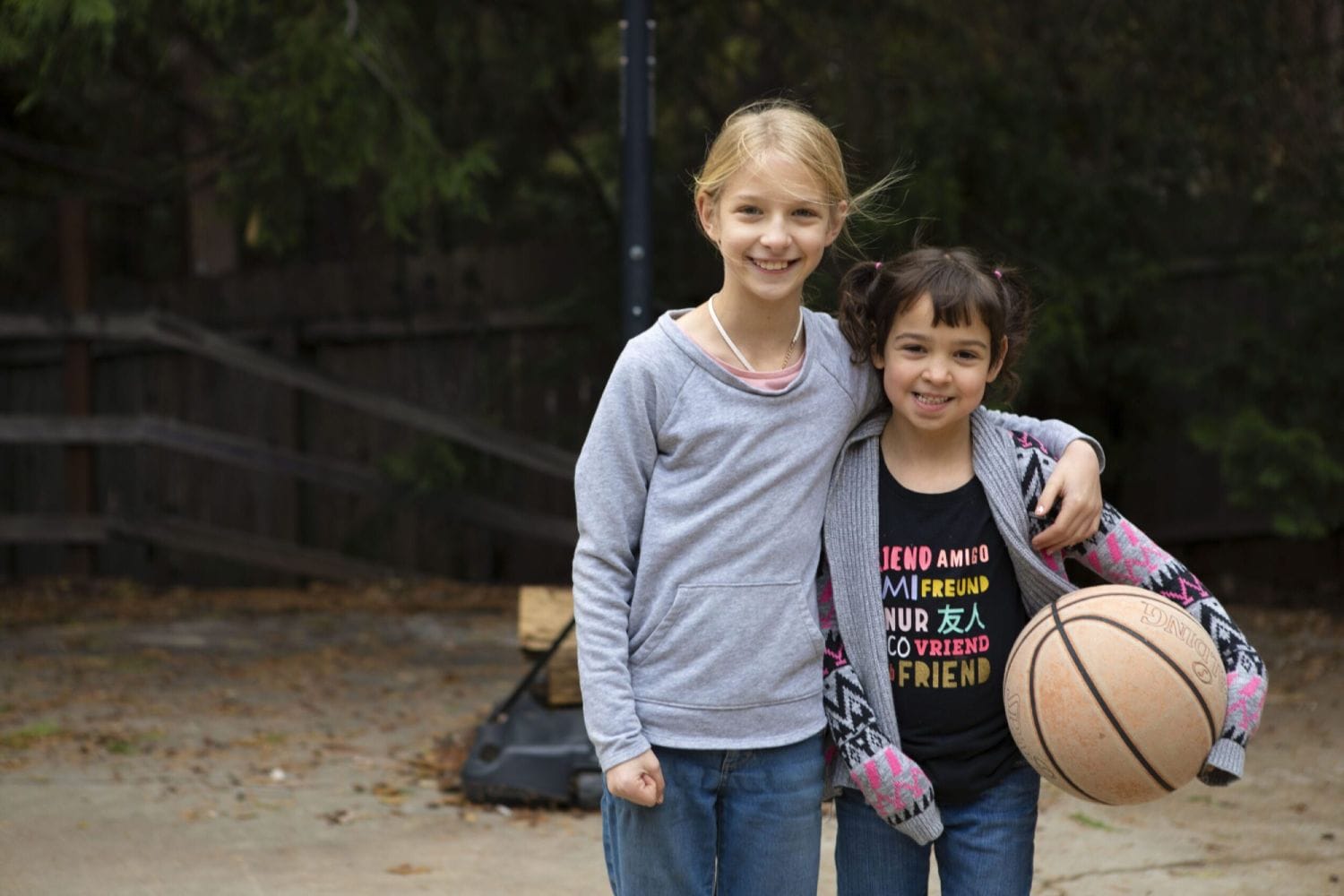 Two young girls smiling outdoors; the taller girl has her arm around the other. The shorter girl holds a basketball. Both wear casual clothes, conveying friendship.